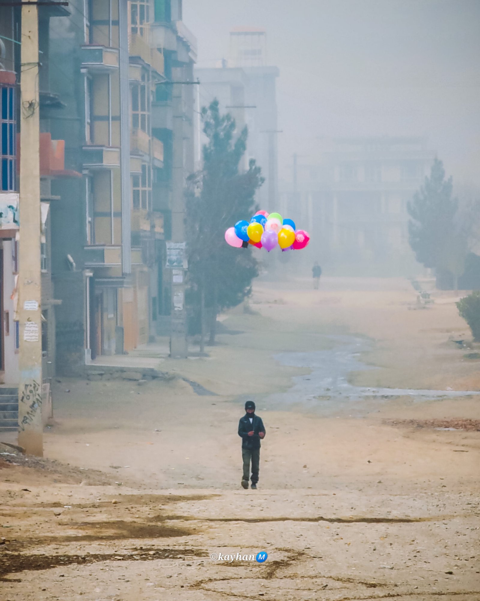 A little boy standing in the middle of the street, holding a bunch of colourful balloons.